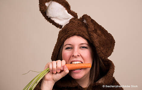 Eine Frau im Hasenkostüm beißt in eine Karotte. Foto: thecherrytree/Adobe Stock.