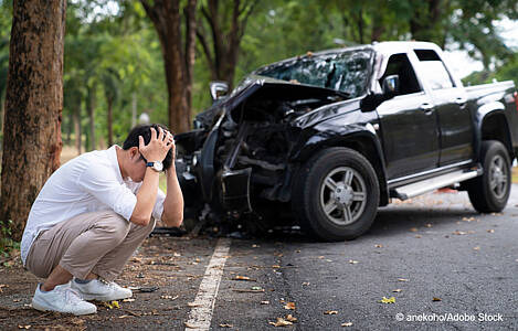 Ein verzweifelter junger Mann hockt vor seinem beschädigten Unfallauto. Foto: anekoho/Adobe Stock.